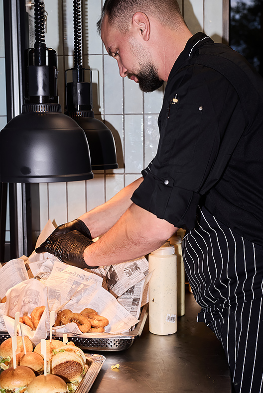 Koch in schwarzer Jacke und gestreifter Schürze richtet mit Handschuhen Zwiebelringe an, daneben stehen Mini-Burger auf einem Tablett.